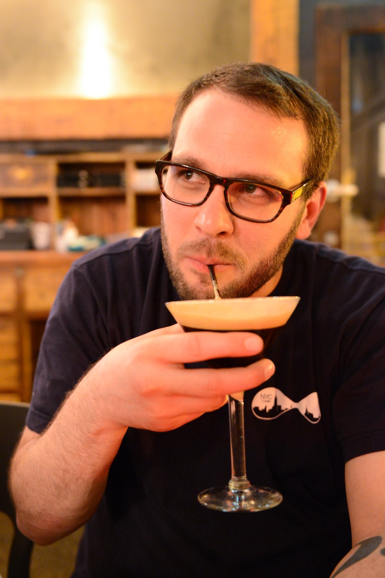 A man wearing glasses sips a cocktail through a straw while seated indoors.
