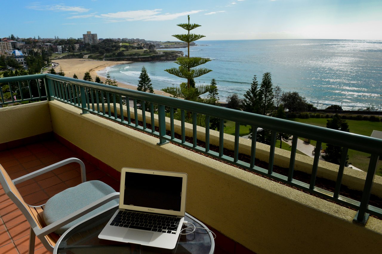 A laptop on a glass table on a balcony overlooking a beach and ocean.