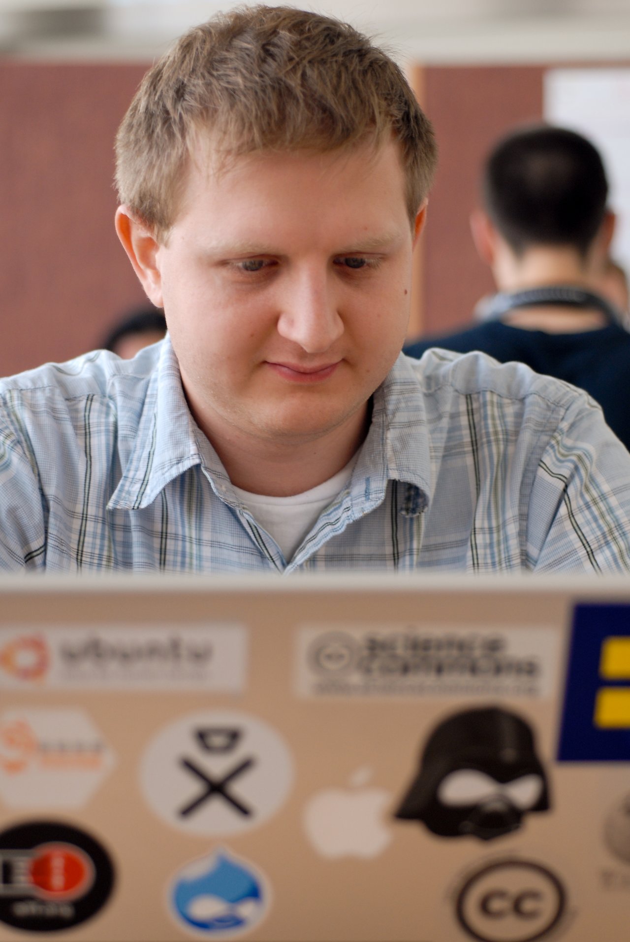 A person in a plaid shirt is focused on a laptop covered in various stickers at a conference.