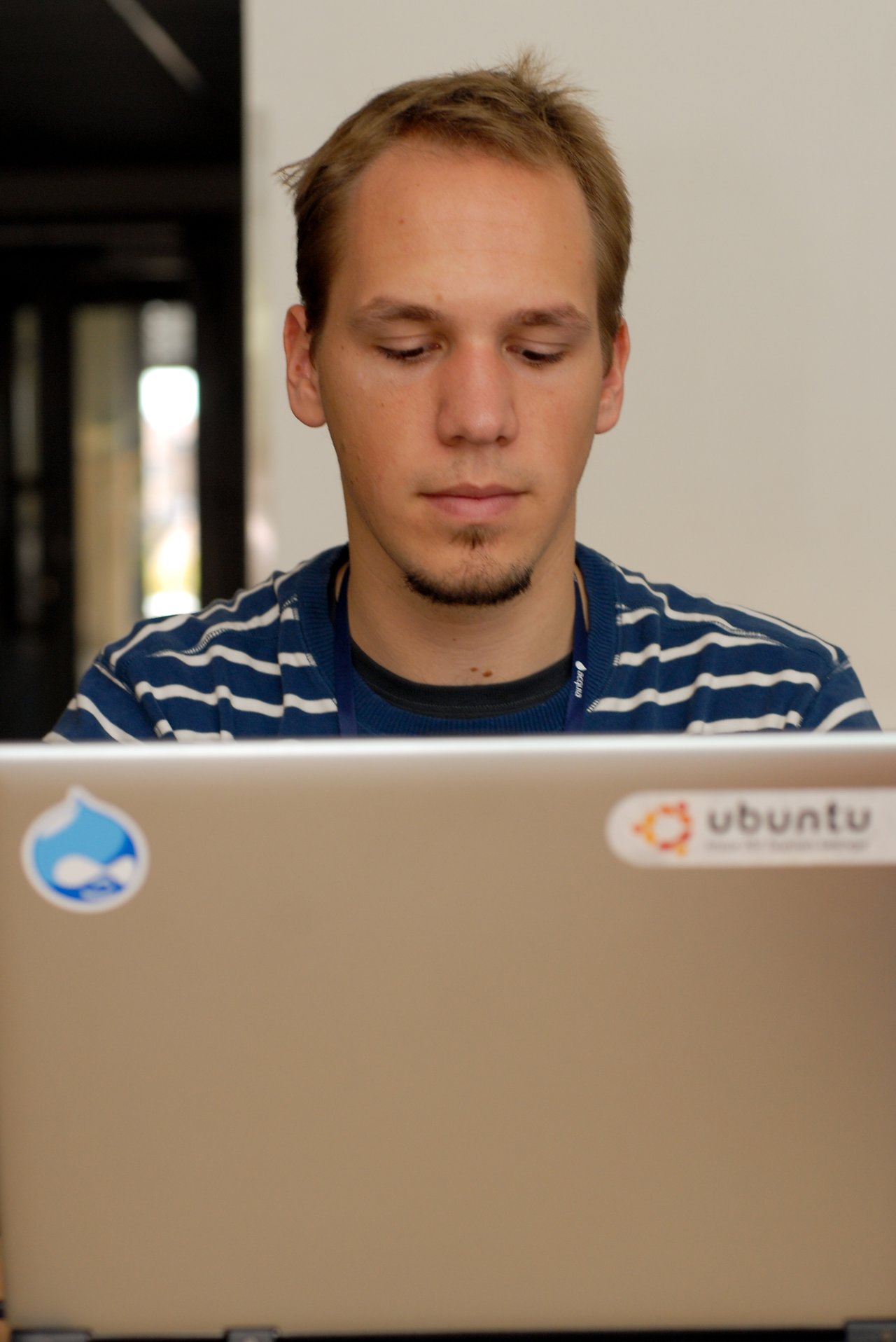 A person in a striped shirt is focused on their laptop, which has Drupal and Ubuntu stickers on it.