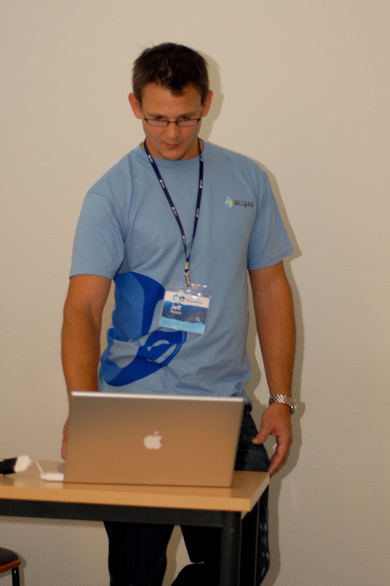 A man in a blue Drupal t-shirt stands at a table, looking at an open MacBook laptop.