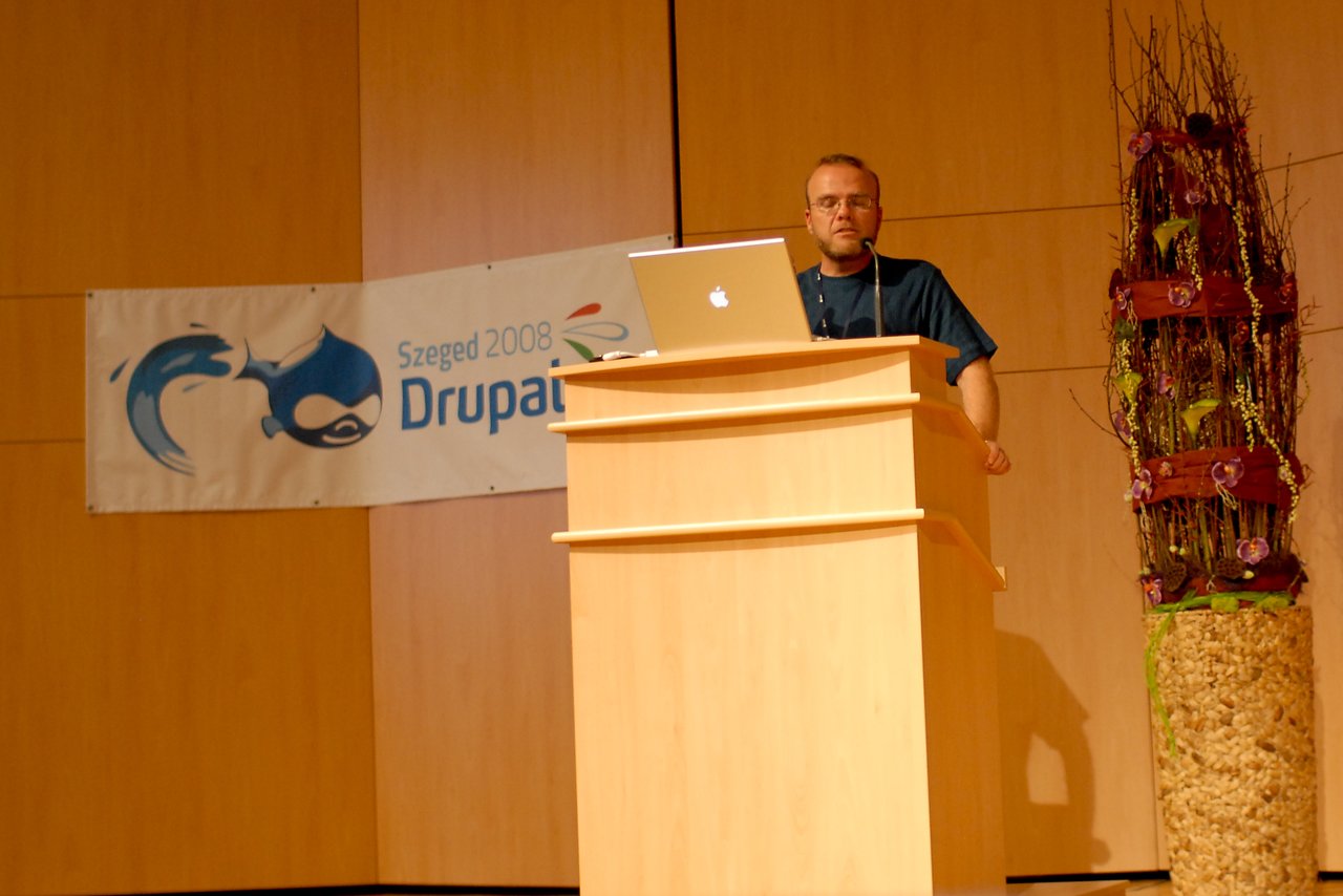 A speaker stands at a podium with a laptop, presenting at DrupalCon Szeged 2008.