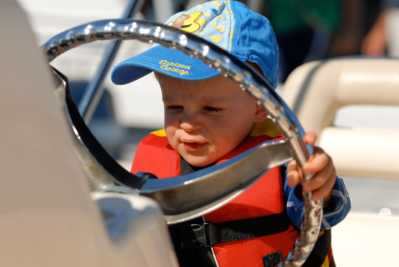 A young child wearing a life jacket and cap grips a boat's steering wheel with a focused expression.