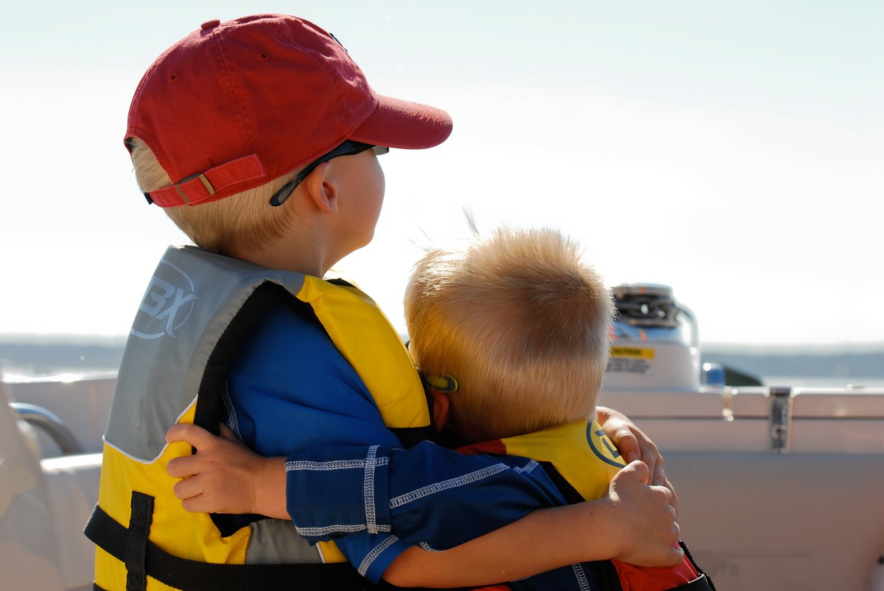 Two young children wearing life jackets sit on a boat, with one hugging the other while looking at the water.