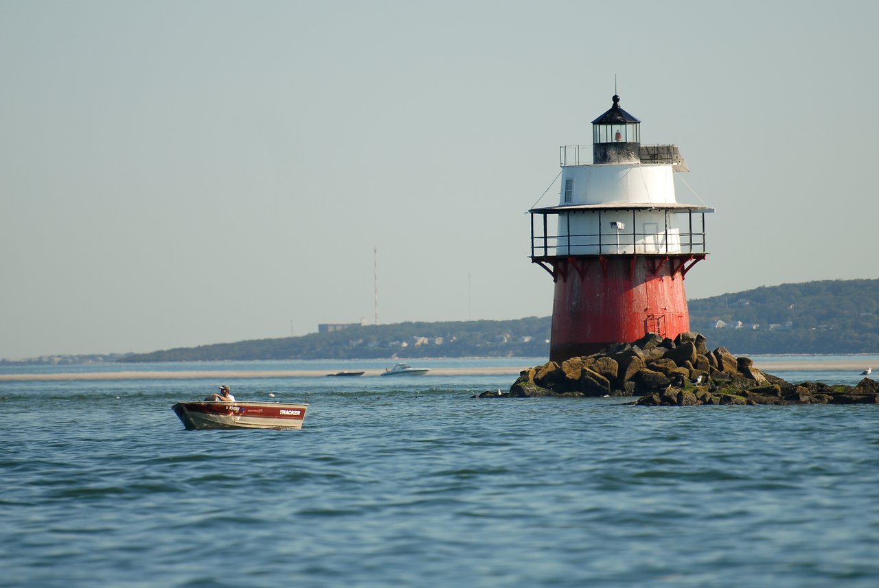 A small boat with a person onboard floats near a red and white lighthouse on a rocky outcrop.