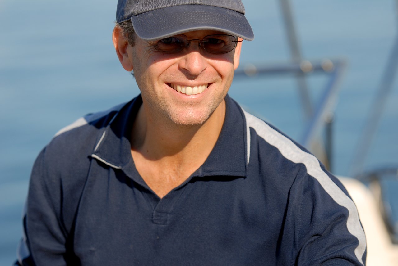 A man wearing sunglasses and a cap smiles while sitting on a boat in sunny weather.