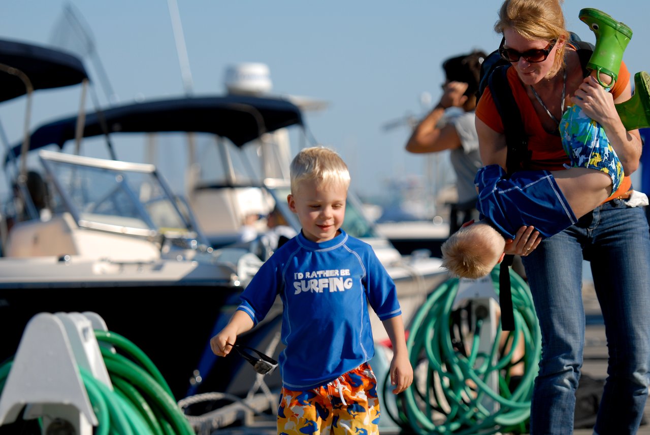 A young boy walks on a dock smiling, while a woman carries another child upside down in her arms.