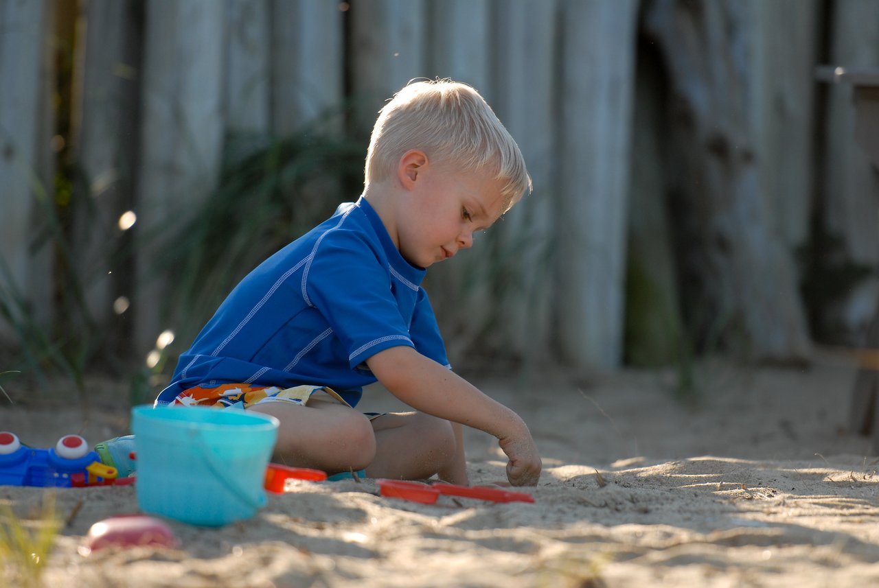 A young child in a blue shirt plays in the sand with a shovel and bucket.