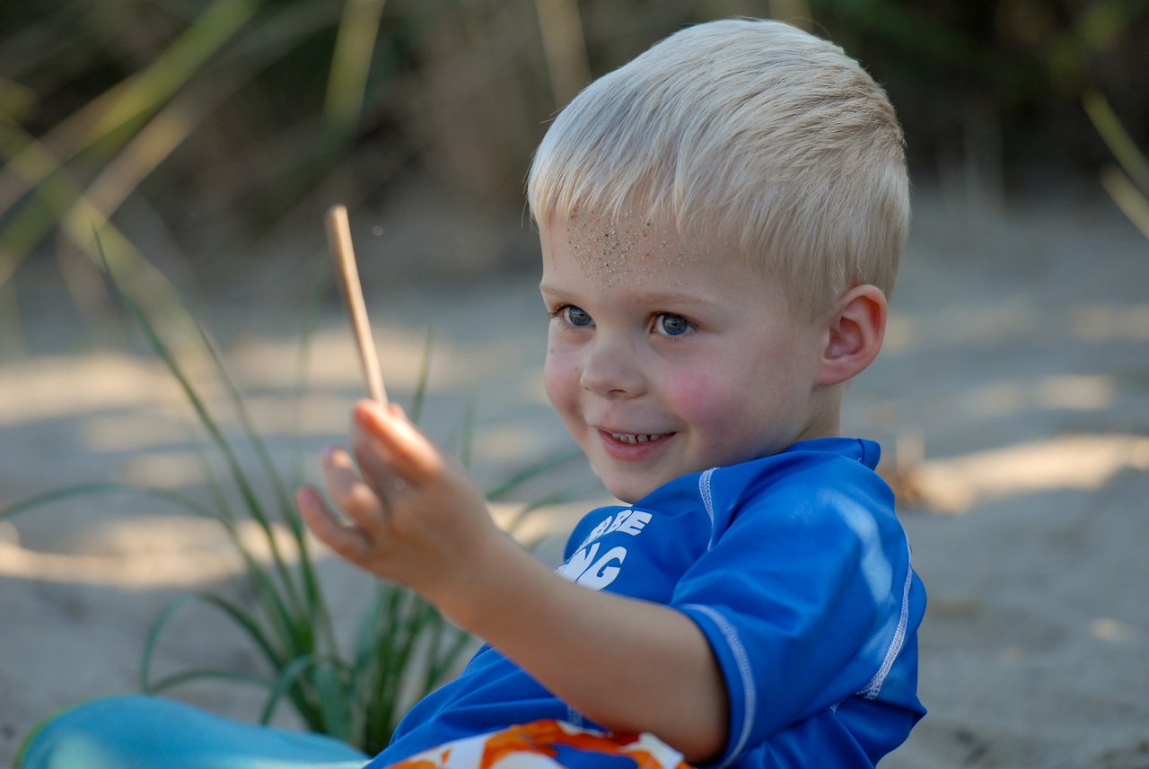 A young child in a blue shirt sits on the sand, smiling and holding a small stick.