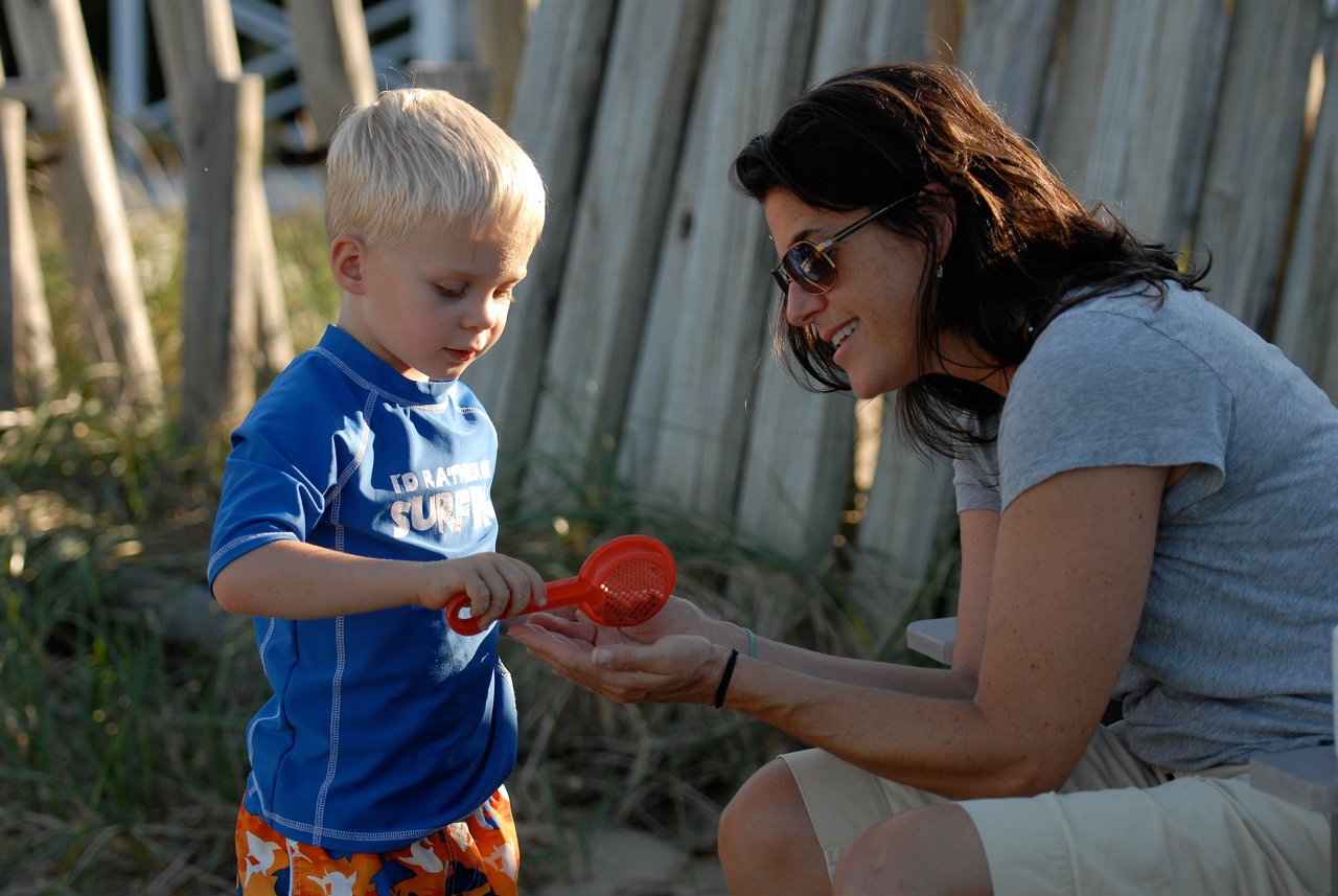 A young boy in a blue shirt pours sand through a small red sieve while a woman watches and smiles.