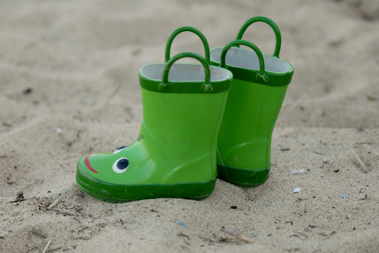 A pair of green children's rain boots with frog faces sit on sandy ground.