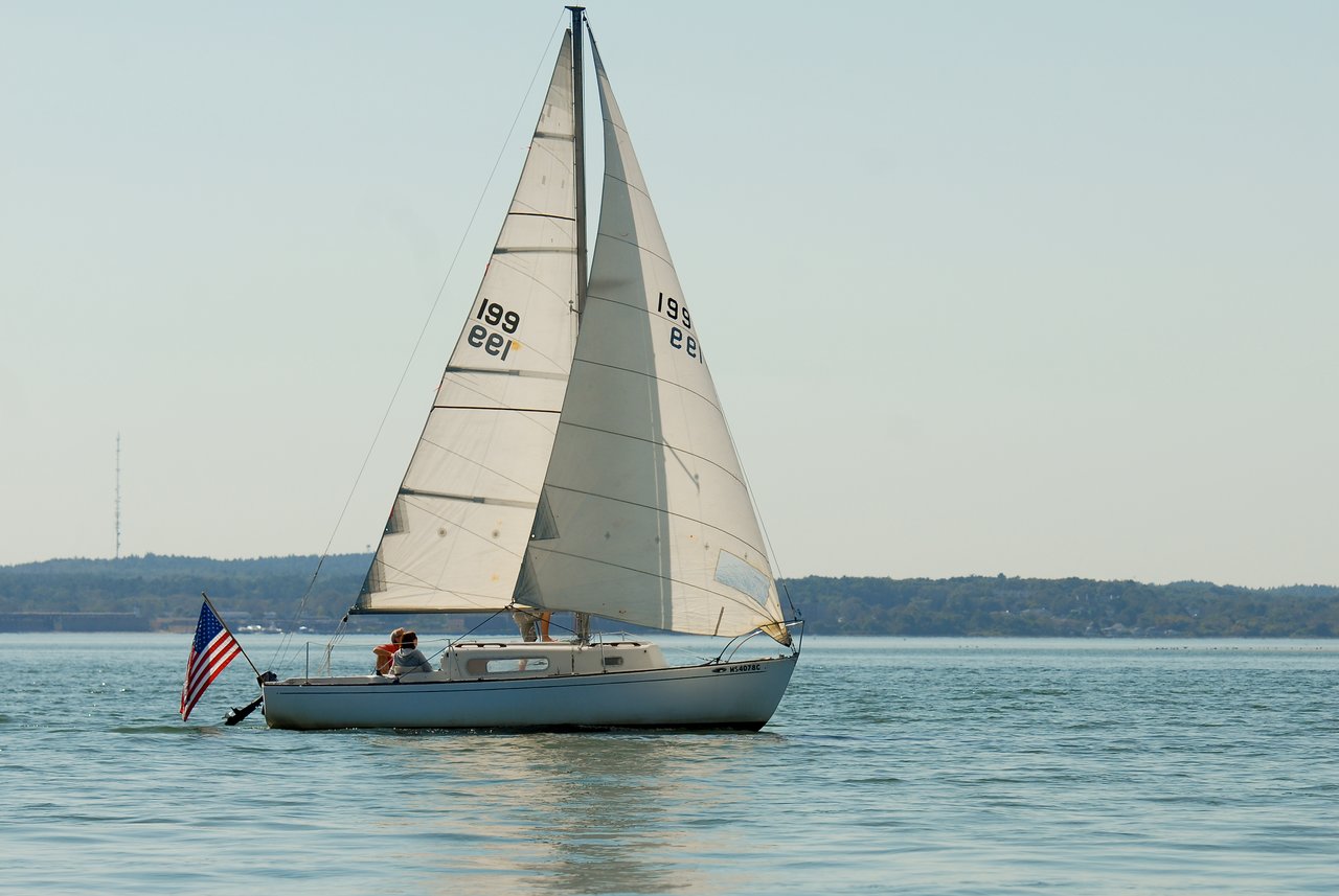 A sailboat with two people on board moves across calm water, with an American flag at the stern.