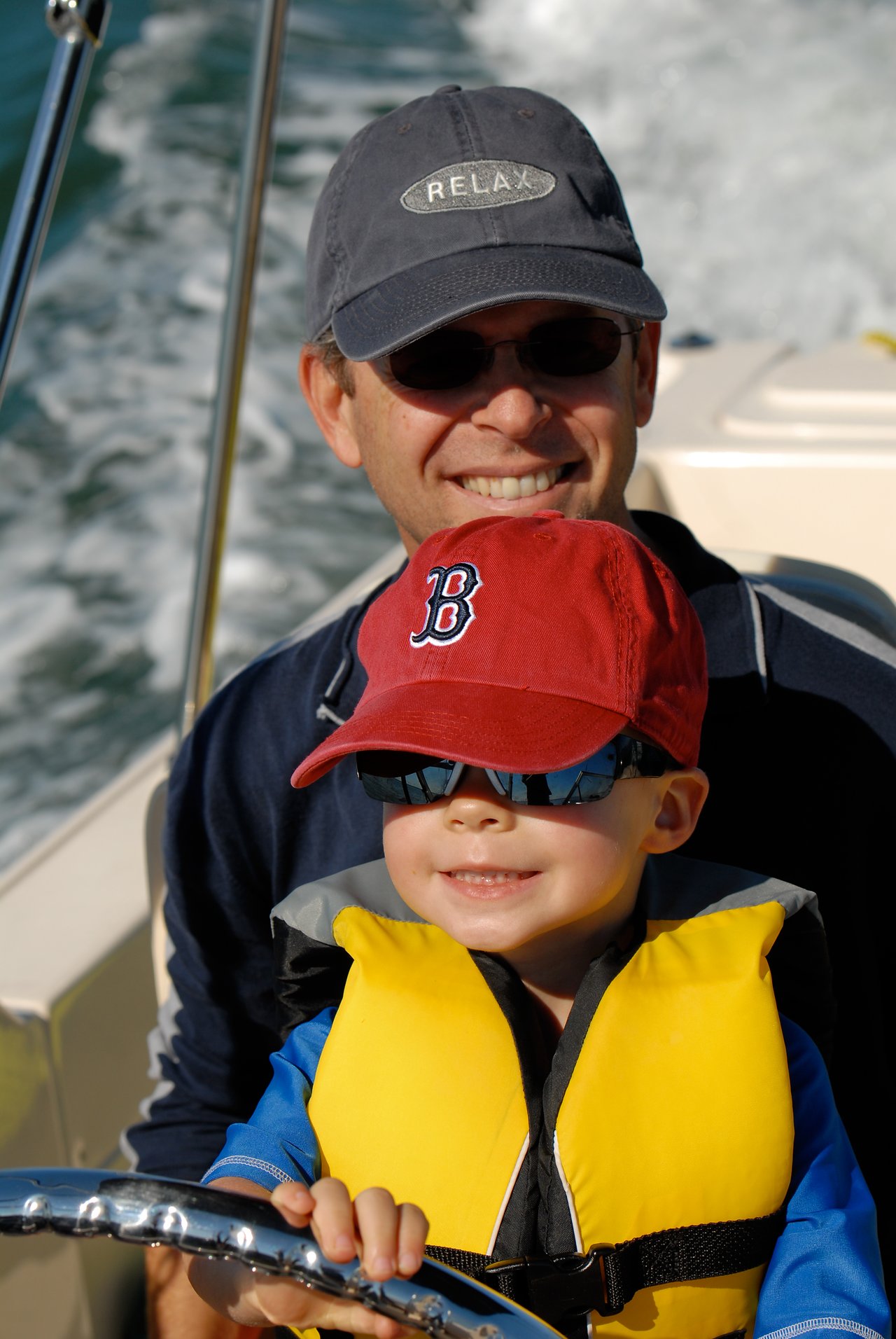A child wearing a life jacket and sunglasses steers a boat, with an adult smiling behind them.