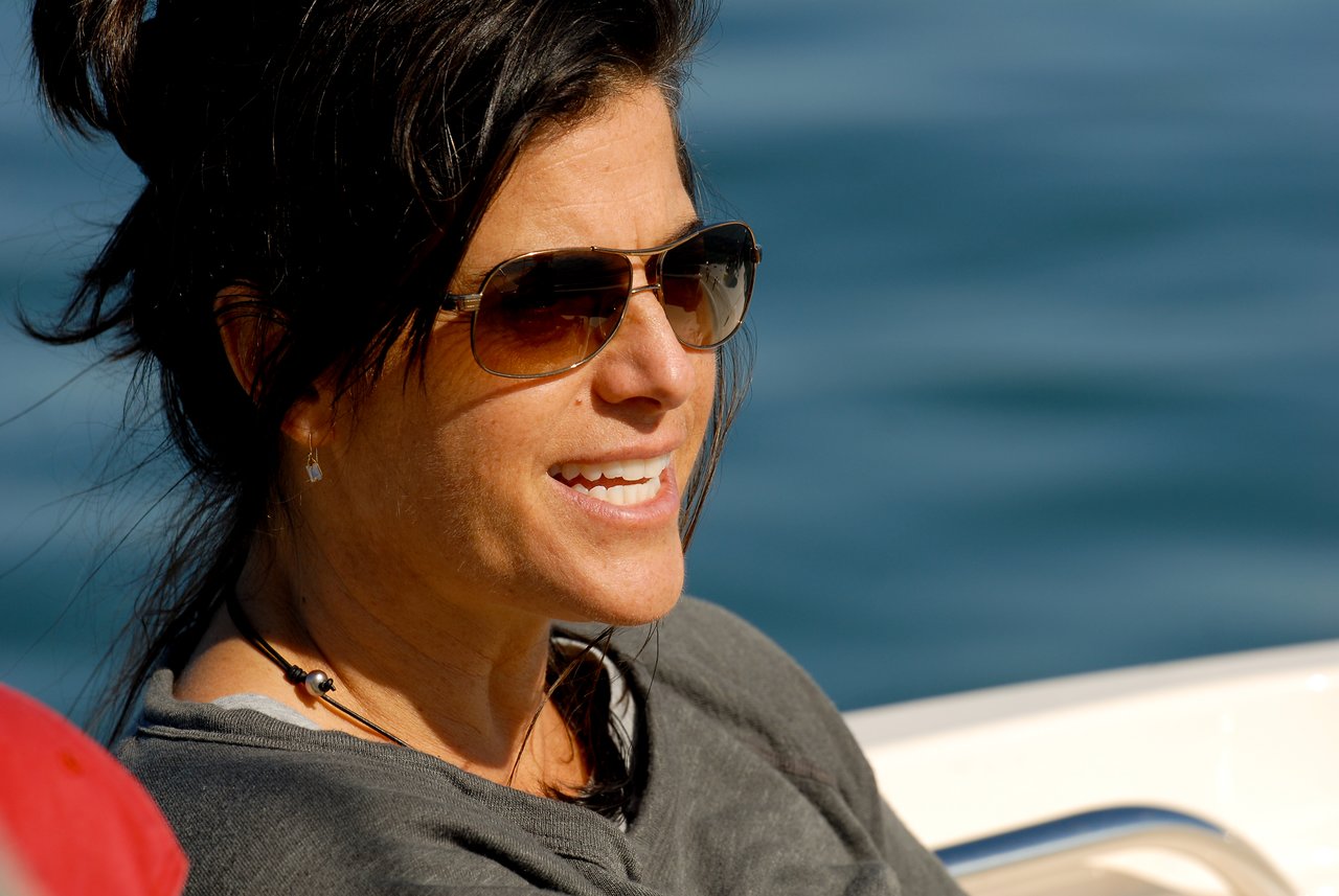 A woman wearing sunglasses and a dark top smiles while sitting on a boat with water in the background.