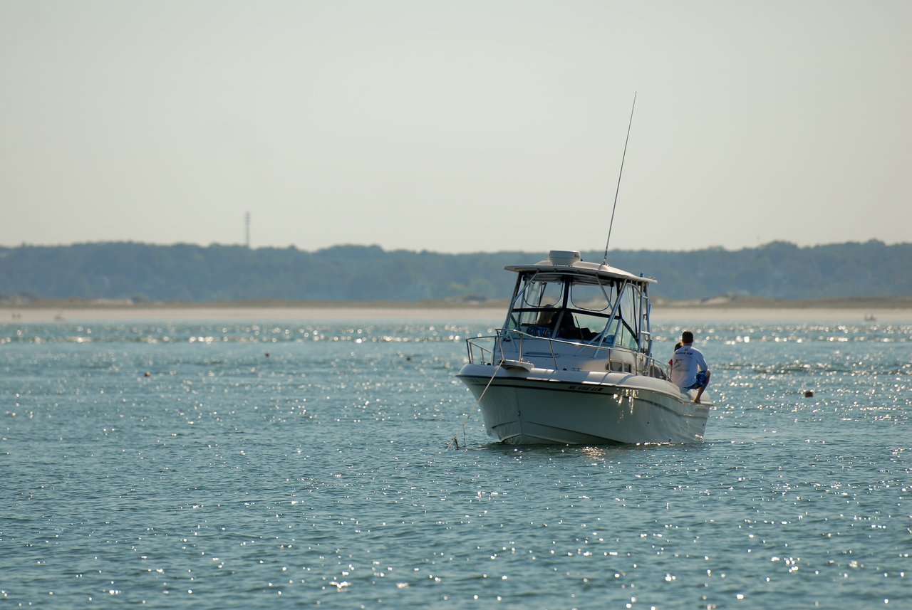 A person sits on the edge of a small motorboat floating on calm water near the shore.