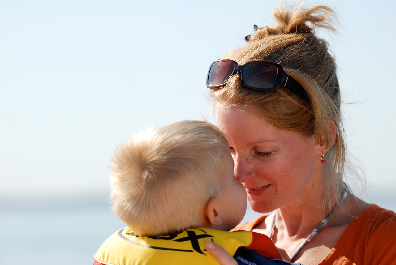 A woman smiles while holding a young child wearing a life jacket, their faces close together.