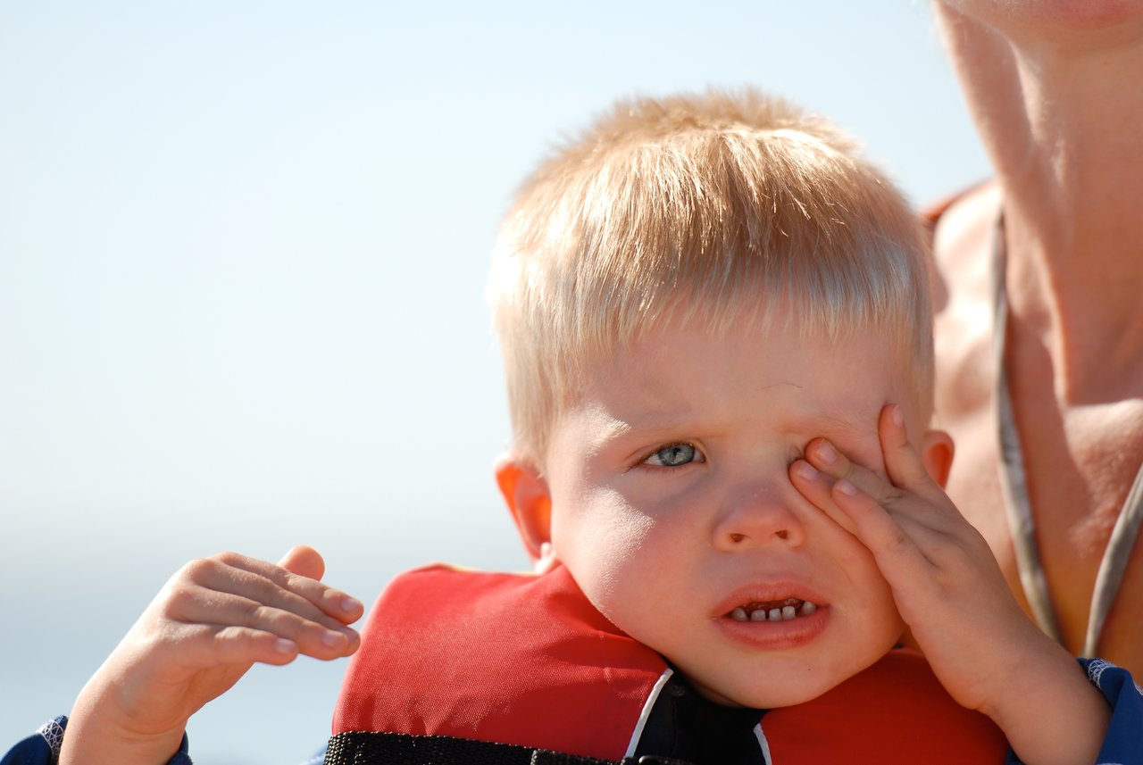 A young child in a life jacket rubs their eye while sitting with an adult in bright sunlight.
