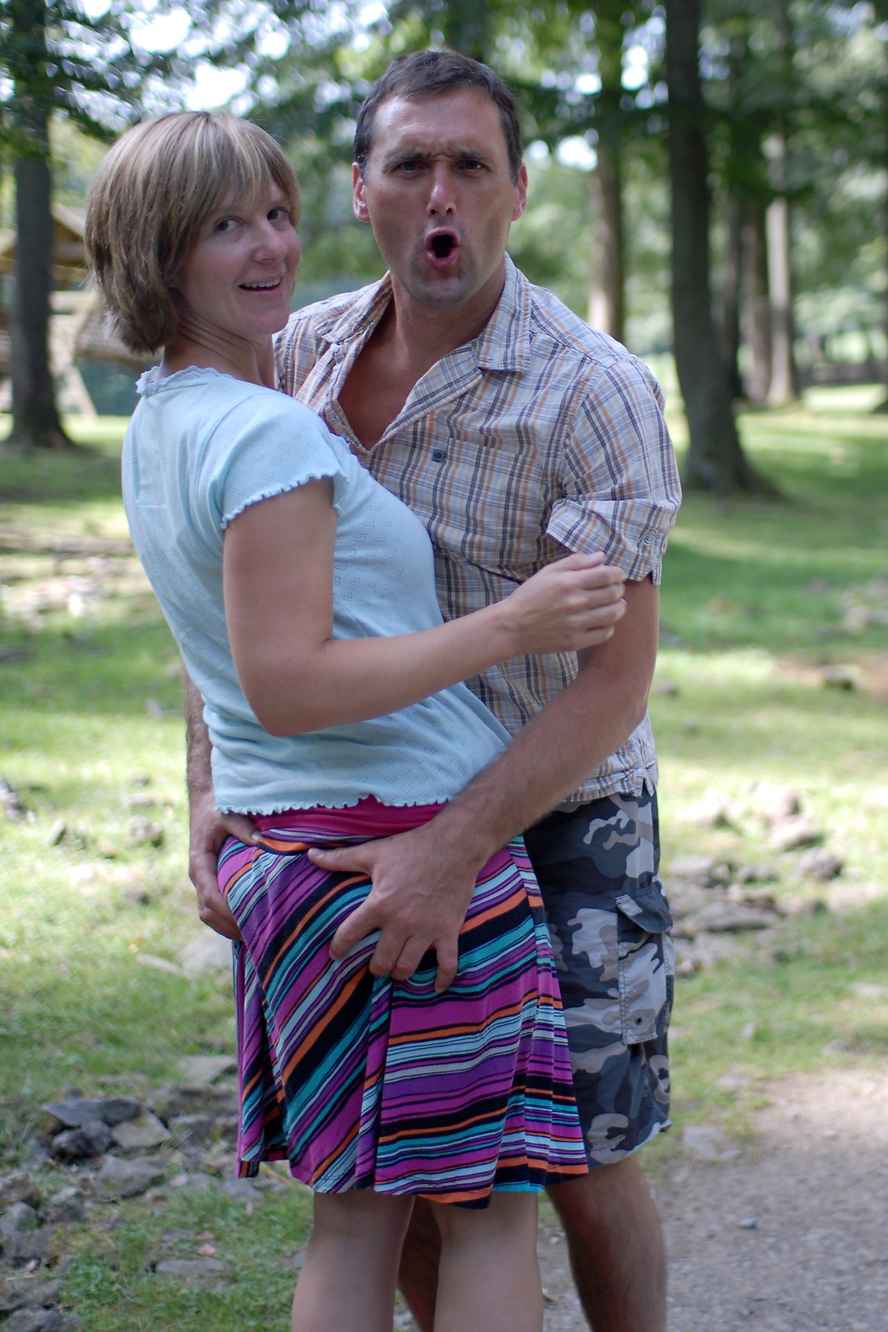A man and woman pose playfully outdoors; the man has an exaggerated facial expression and his hand on her skirt.