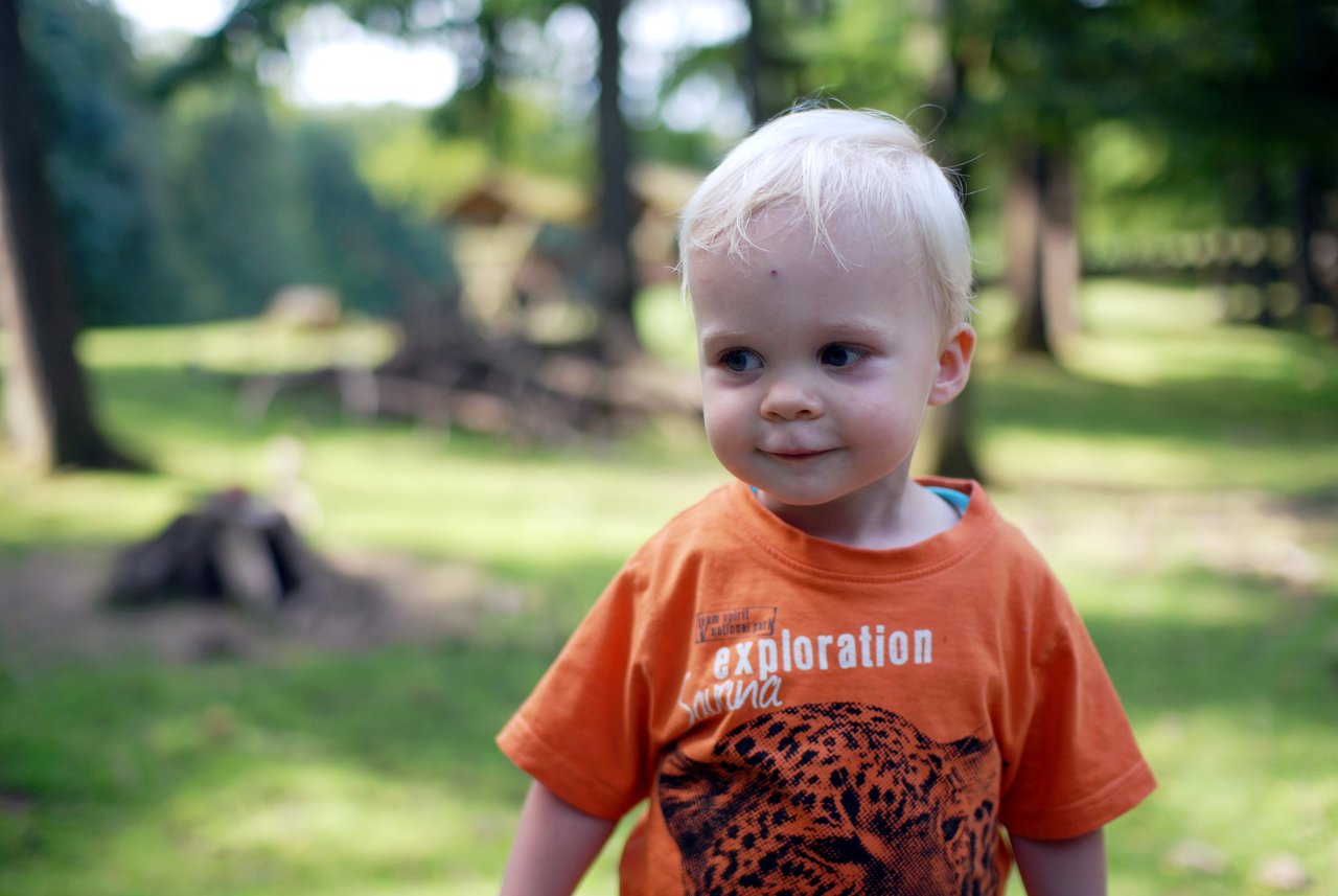 A young child with blonde hair and an orange shirt stands outside, looking to the side with a slight smile.