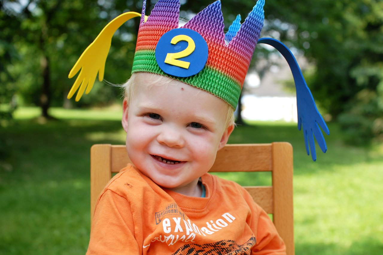 A smiling toddler wearing a colorful birthday crown with the number two sits on a wooden chair outdoors.