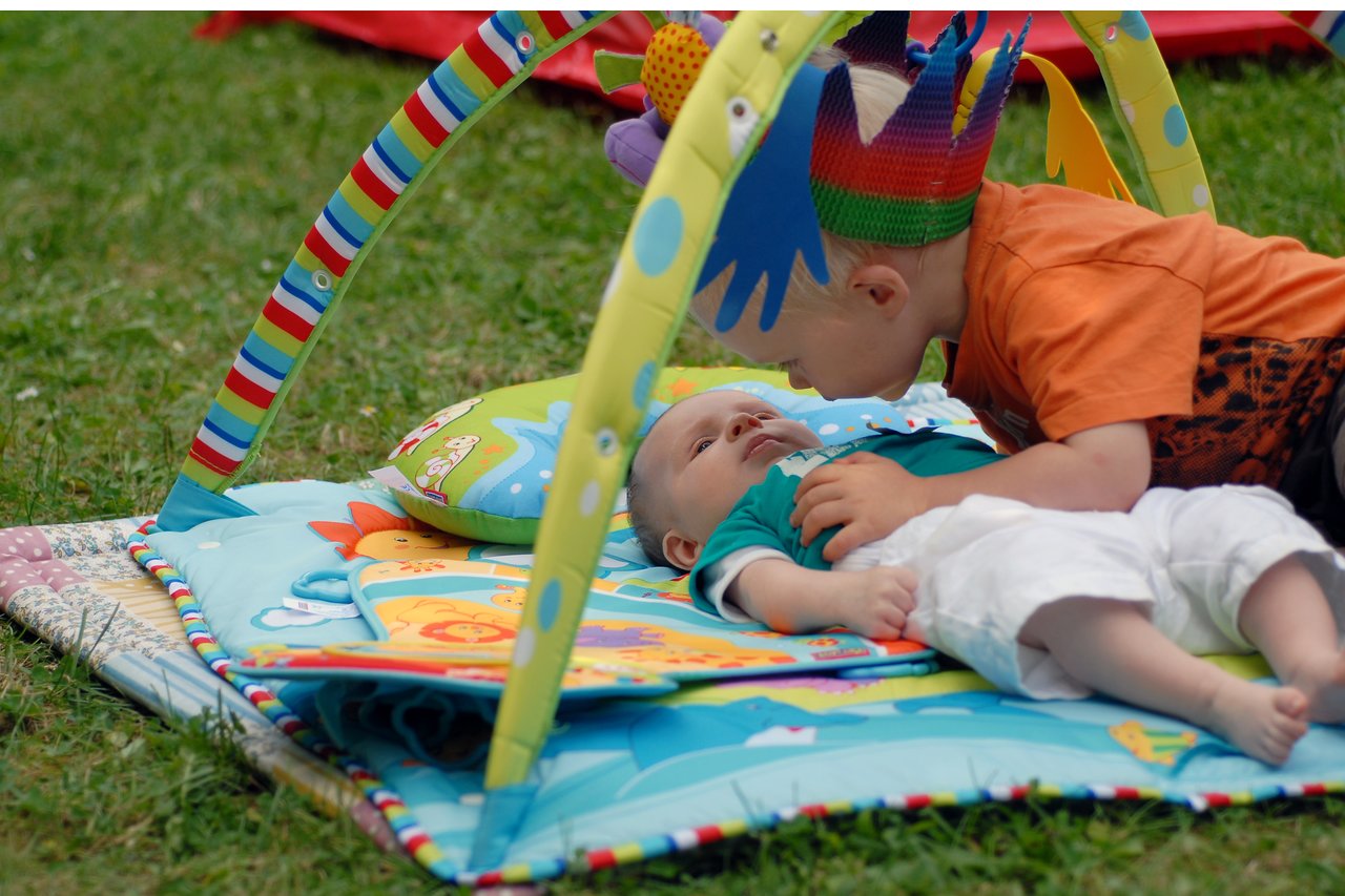A young child wearing a colorful crown leans in close to an infant lying on a play mat outside.
