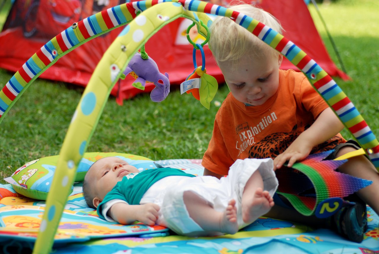 A baby lies on a play mat while a toddler sits nearby, interacting with a colorful toy.