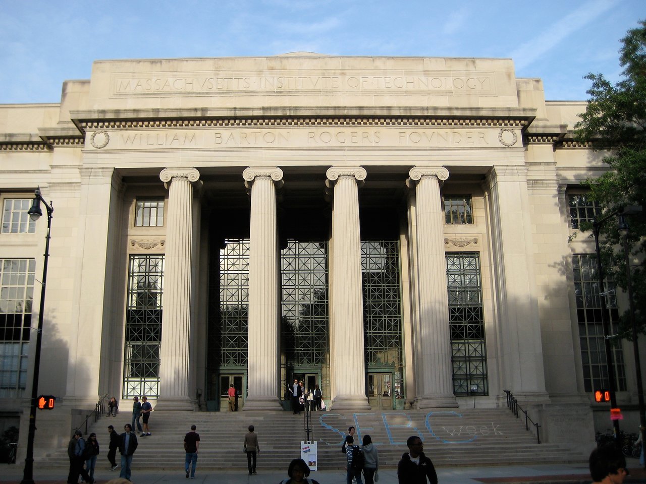 The entrance of an MIT building with tall columns, large windows, and people walking up and down the steps.