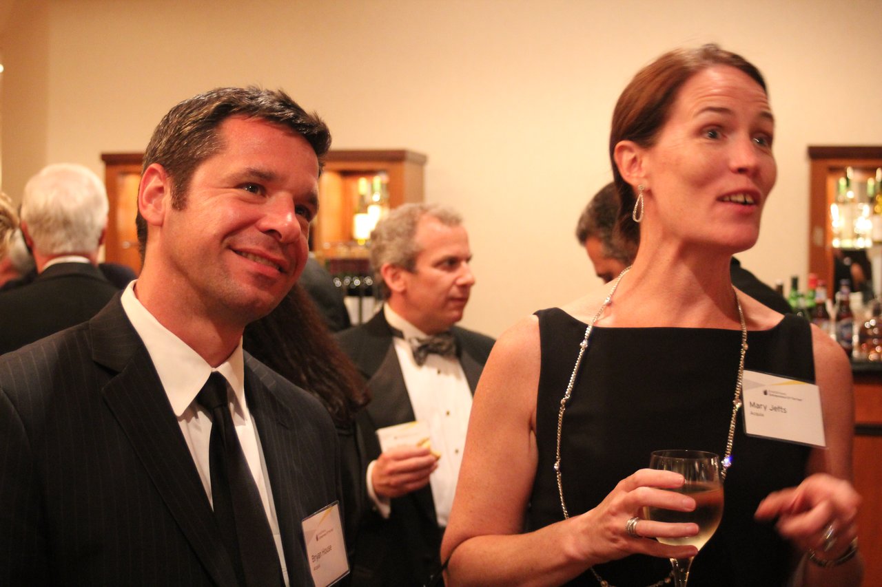 Two people in formal attire converse at an award banquet, with one holding a glass of wine.