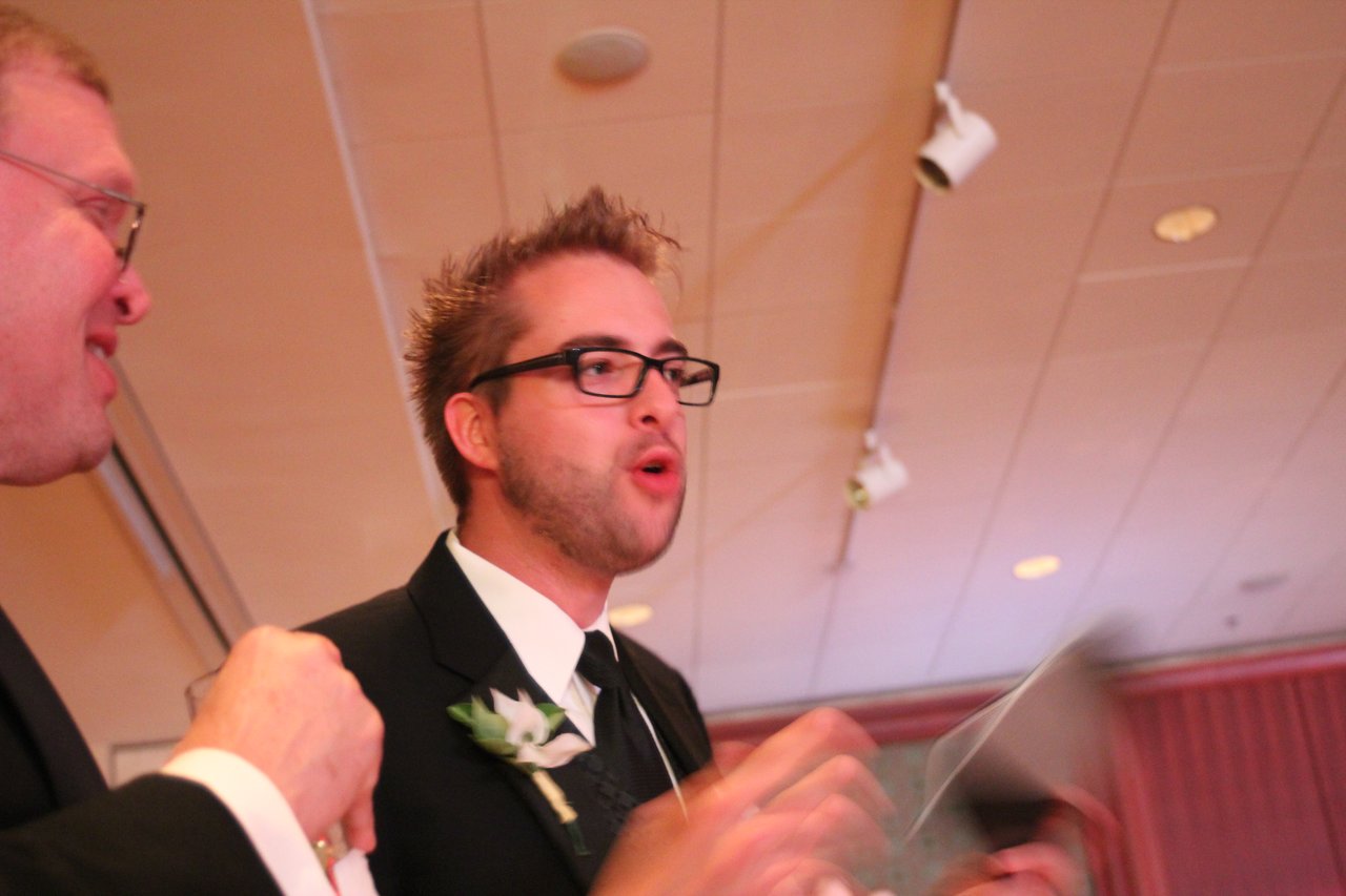 A man in a suit with a boutonniere speaks while holding an award at a banquet.