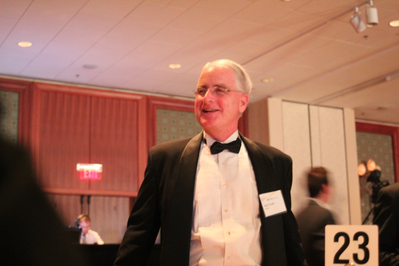 A man in a tuxedo smiles while standing at an award banquet, with other attendees in the background.