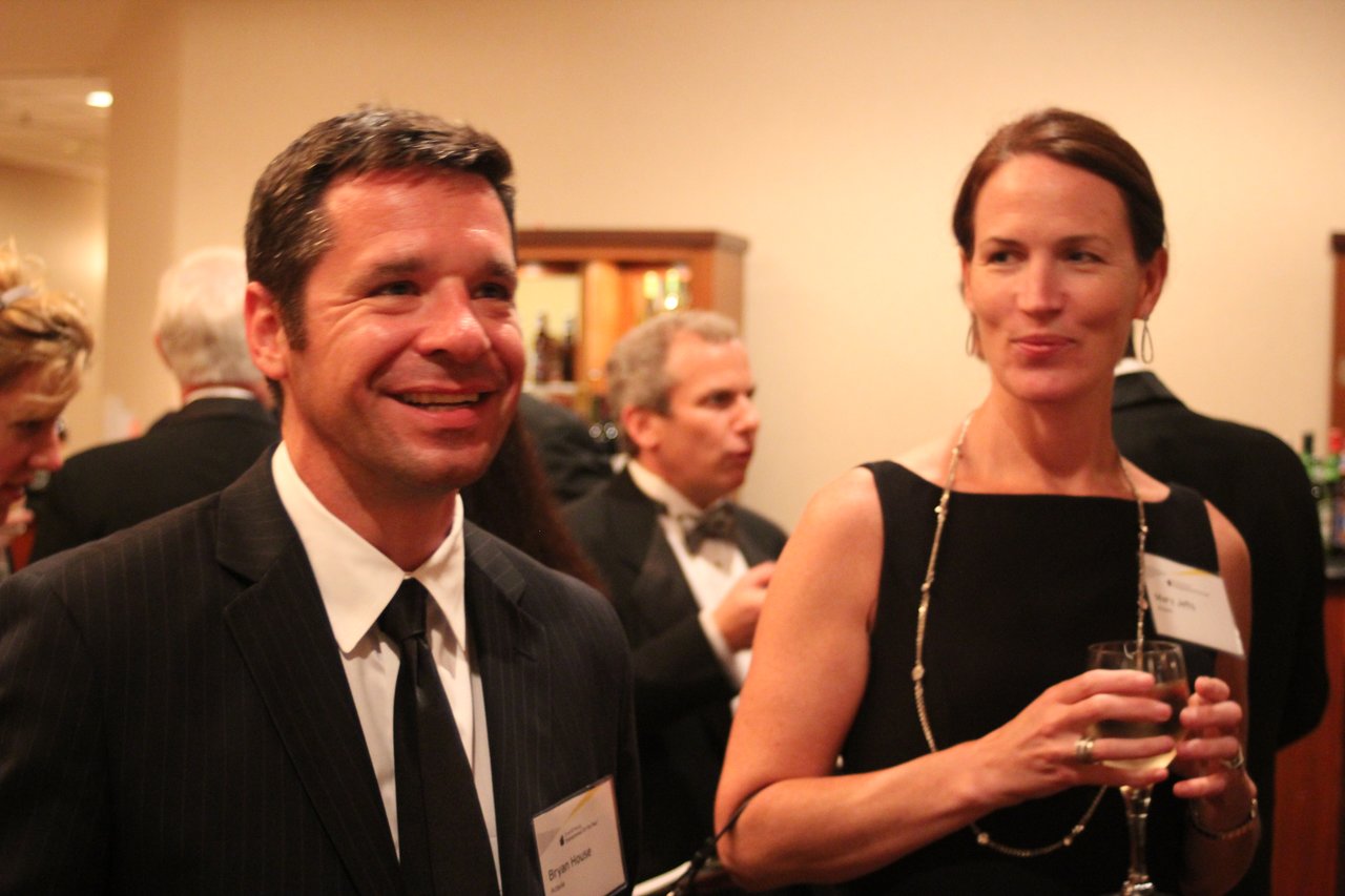 Two people in formal attire smile and converse at an award banquet, holding drinks.