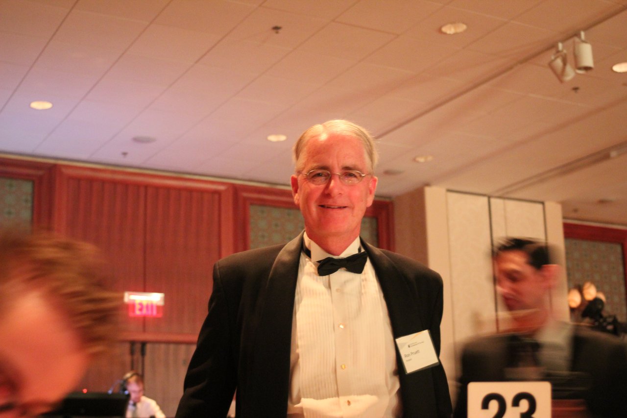 A man in a tuxedo smiles at the camera during an award banquet, with other attendees in the background.