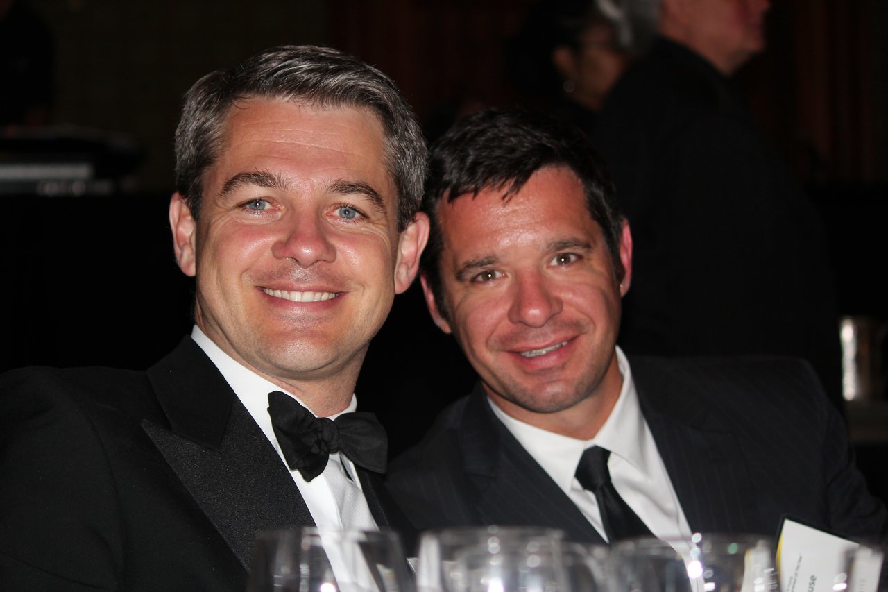 Two men in formal attire smile while seated at a banquet table during an award event.
