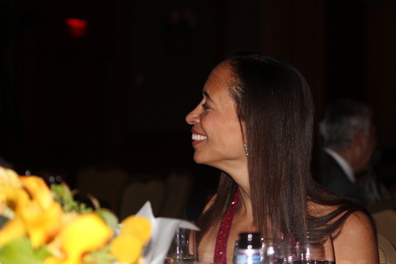 A woman in a red dress smiles while seated at an award banquet table with flowers and drinks.