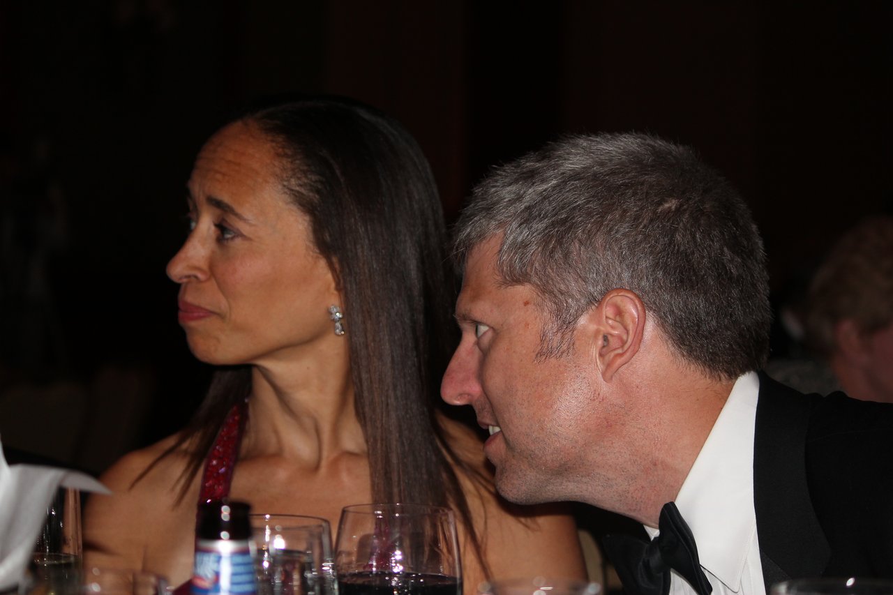 A man in a tuxedo leans in to speak to a woman in a red dress at a banquet.