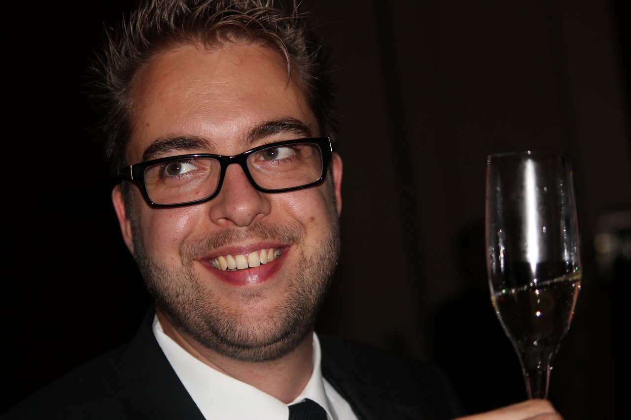A man in a suit and glasses smiles while holding up a champagne glass at an award banquet.