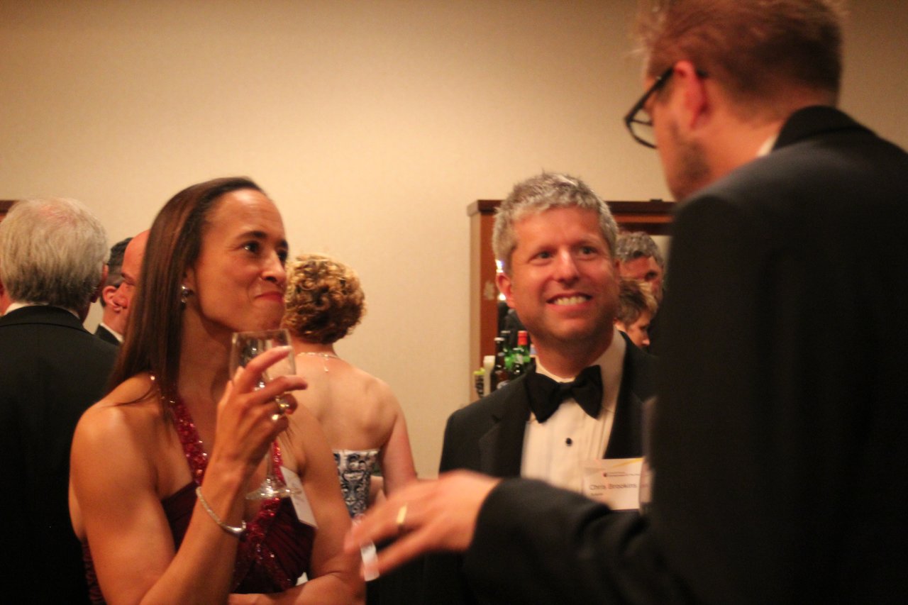 Guests in formal attire converse at an award banquet, with one woman holding a glass and smiling.
