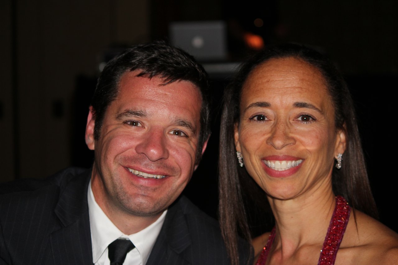 A man in a suit and a woman in a red dress smile at the camera during an award banquet.
