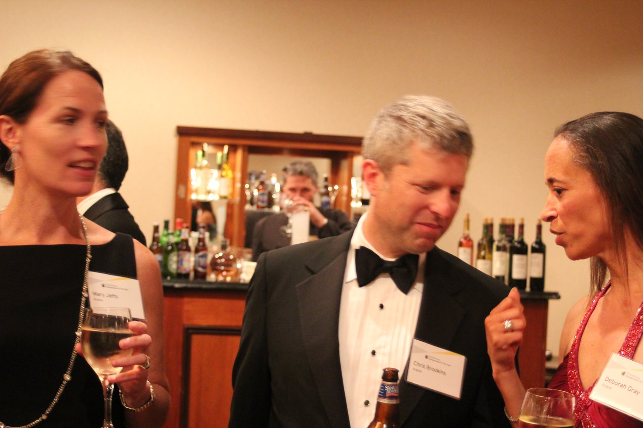 Three people in formal attire converse while holding drinks at an award banquet, with a bar in the background.