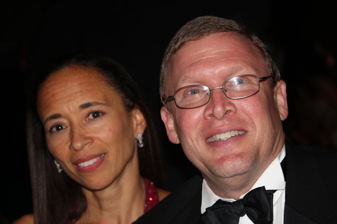 A man in a tuxedo and a woman in a red dress smile at the camera during an award banquet.