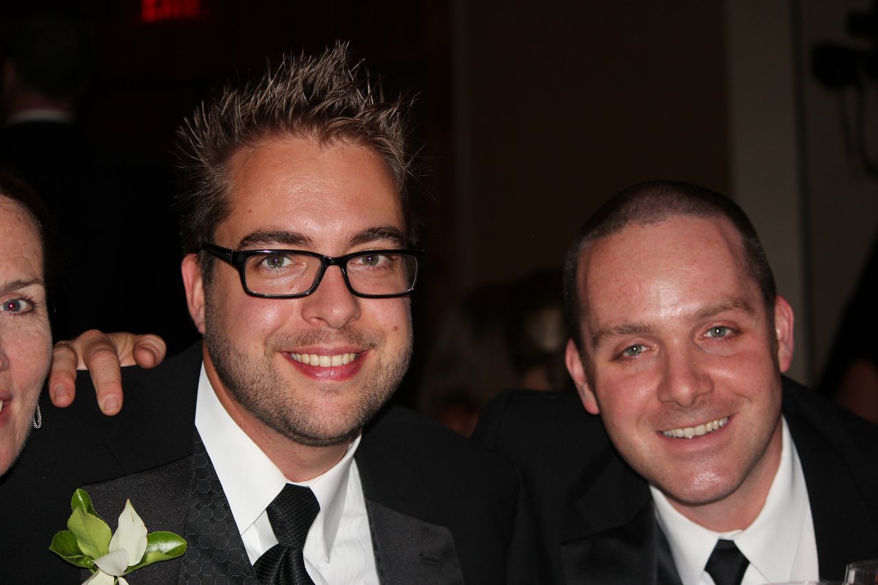 Two men in suits smiling at the camera during an award banquet, with a partial view of another person.