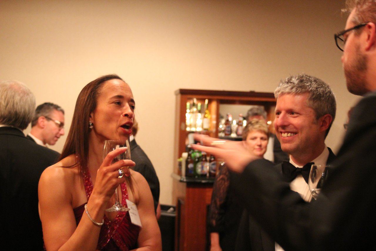 People in formal attire converse and hold drinks at an award banquet, smiling and engaging in discussion.
