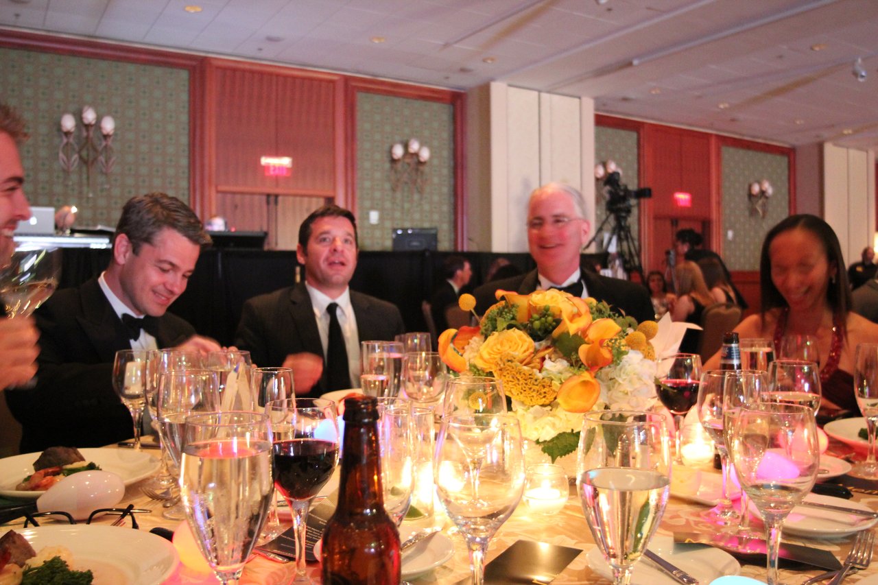 People in formal attire sit around a decorated banquet table, enjoying food, drinks, and conversation at an award event.