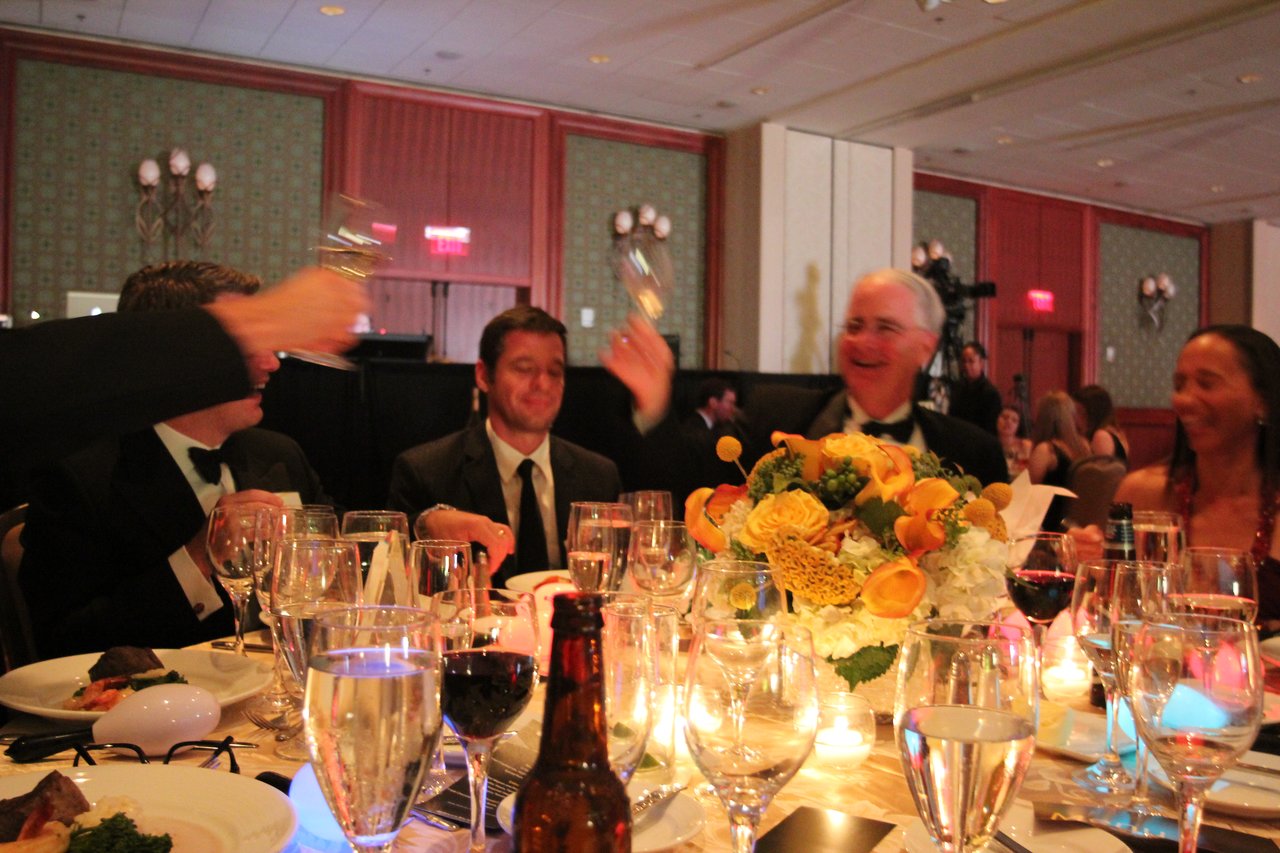 Guests in formal attire raise glasses in a toast at an award banquet, smiling and celebrating together.