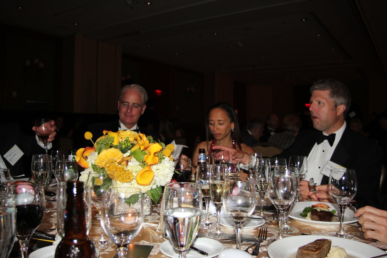 Guests in formal attire sit at a banquet table, eating, drinking, and conversing during an award event.