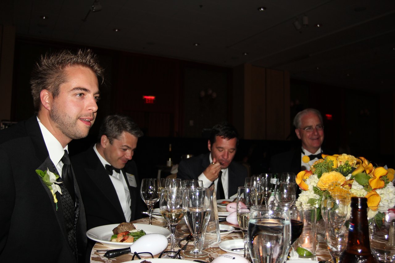 Four men in tuxedos sit at a banquet table, eating and talking, with wine glasses and flowers in front.