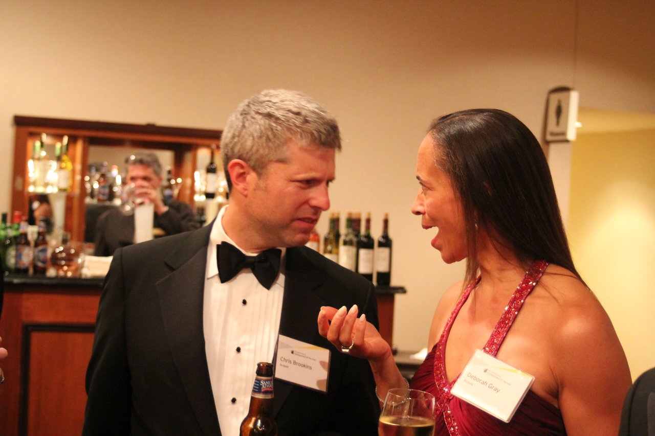A man in a tuxedo and a woman in a red dress are talking at an award banquet, holding drinks.