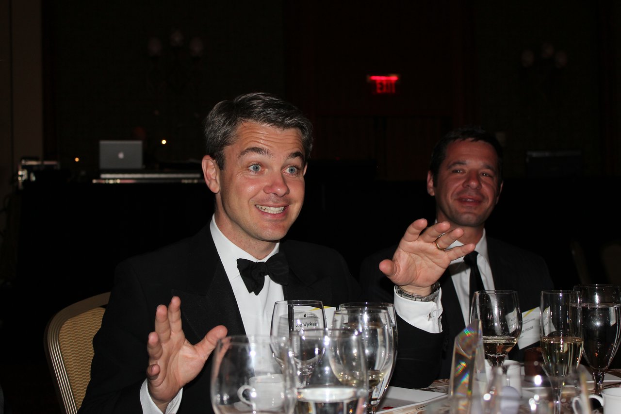 Two men in tuxedos sit at a banquet table, smiling and gesturing while engaged in conversation.