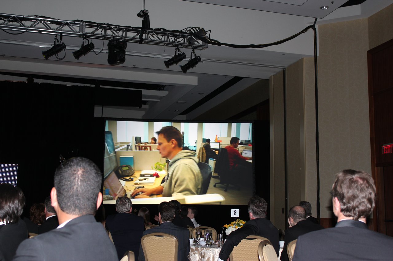 Attendees in formal attire watch a large screen displaying an office scene during an award banquet.