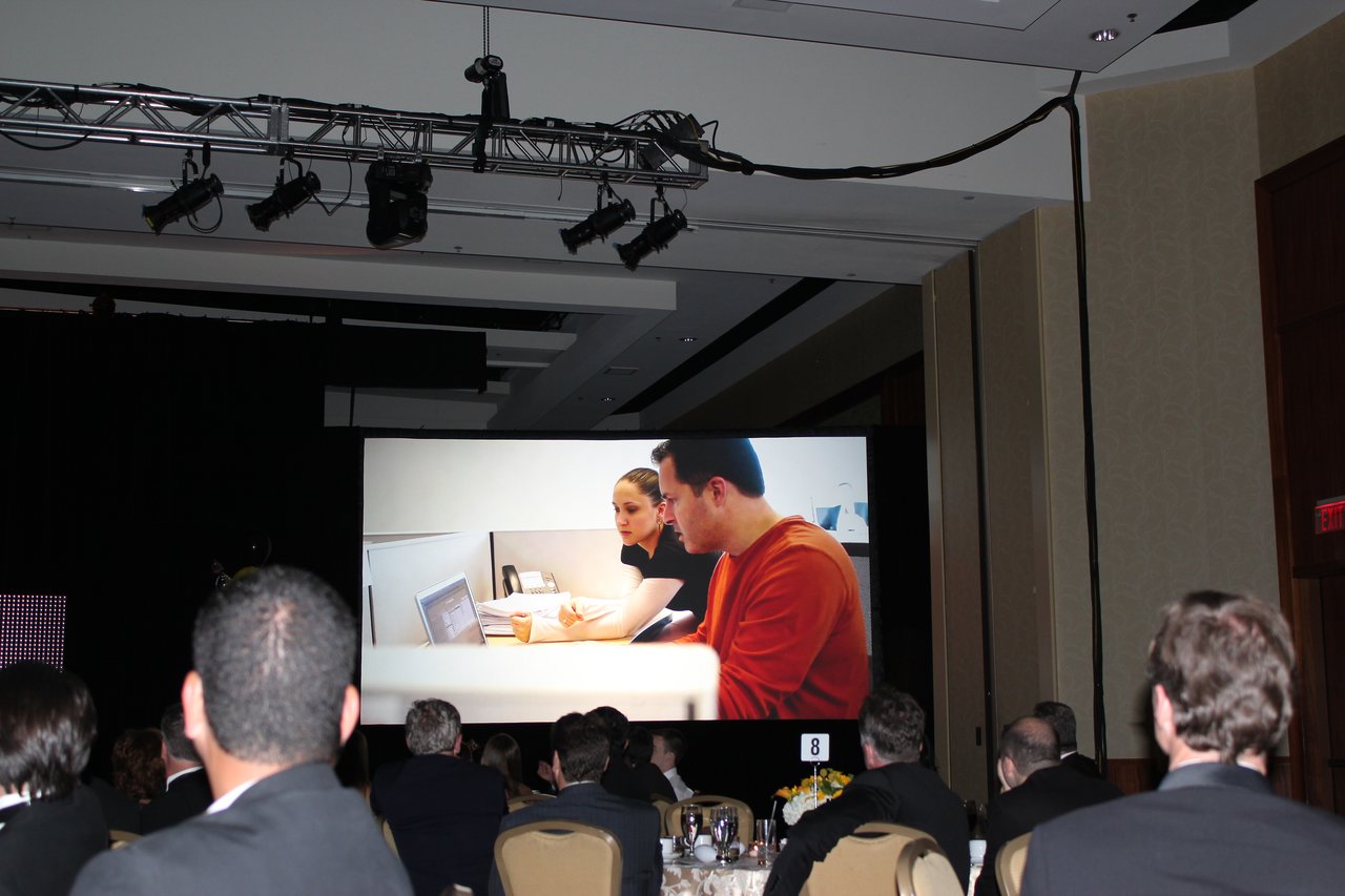 Audience watches a large screen displaying an image of two people working together in an office setting.
